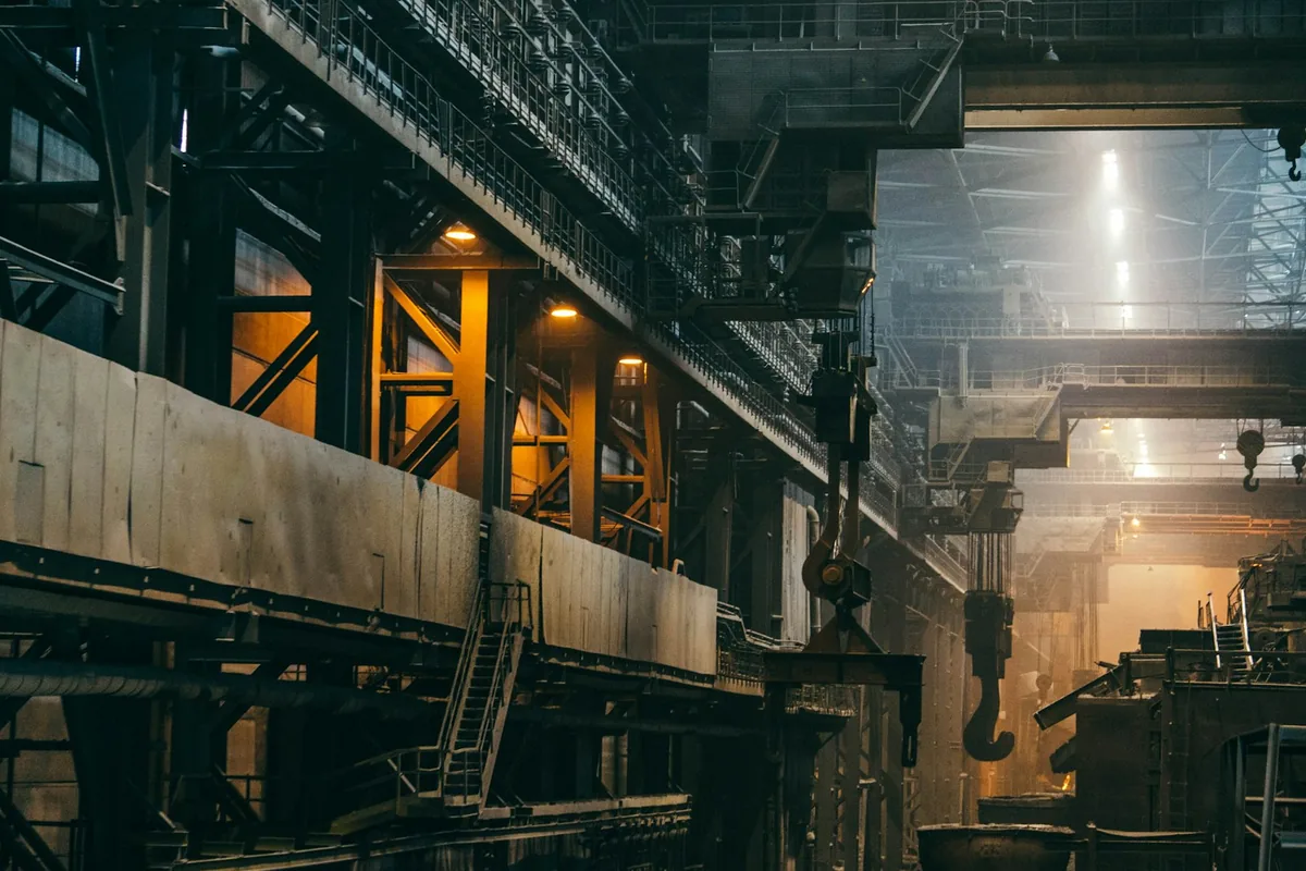 Large industrial steelworks interior with overhead cranes, steel beams and warm light.