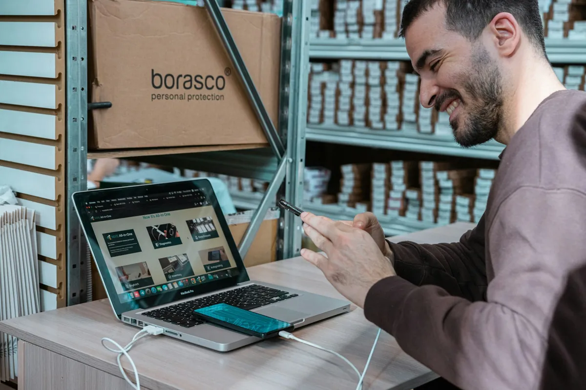 Warehouse professional using a laptop and phone beside inventory shelves