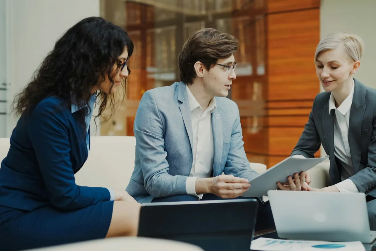 Three business professionals review a folder together beside open laptops.