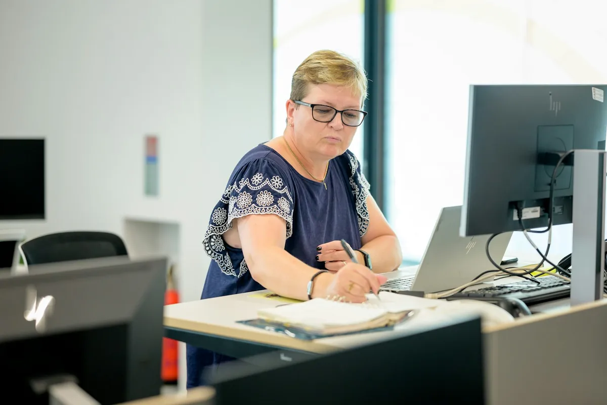 A woman reviews and writes in a document binder beside a laptop and office monitor.