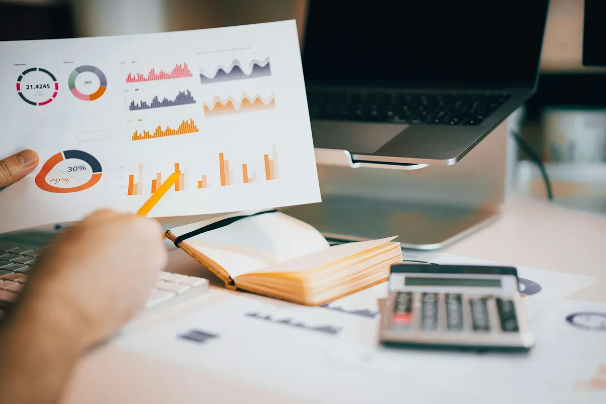 An analyst points to printed performance charts beside a laptop and calculator.