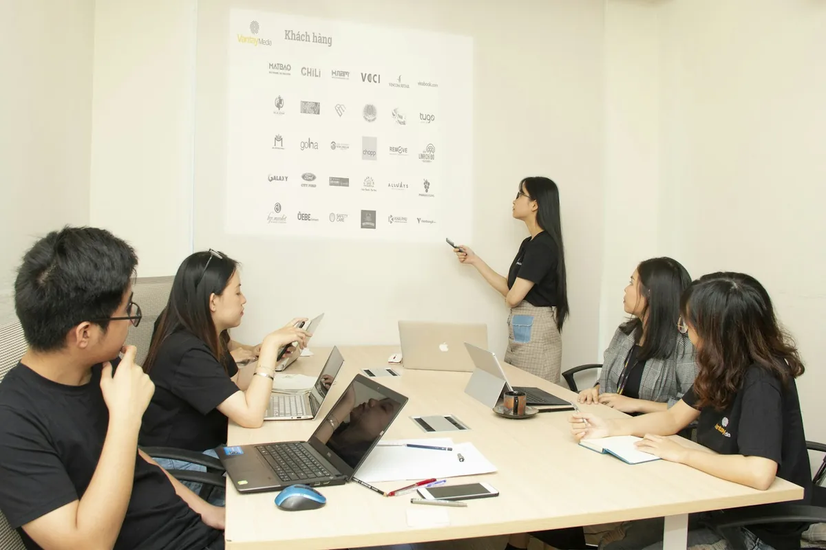 A colleague presents on a large projected screen to a team with laptops and notebooks.
