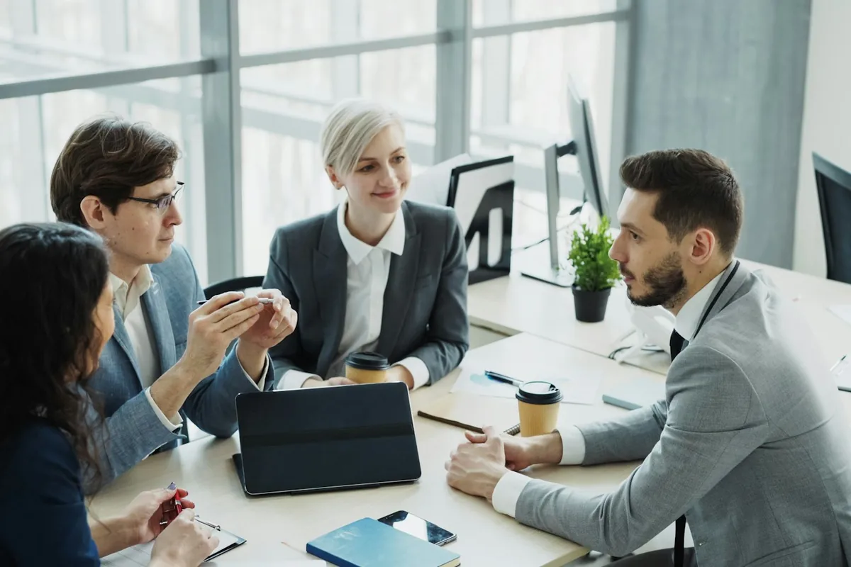 Four business professionals discuss work around a desk with a tablet and printed charts.