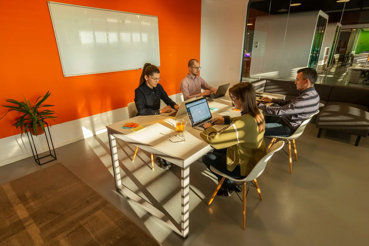 Four colleagues work together on laptops around a meeting table with sticky notes.