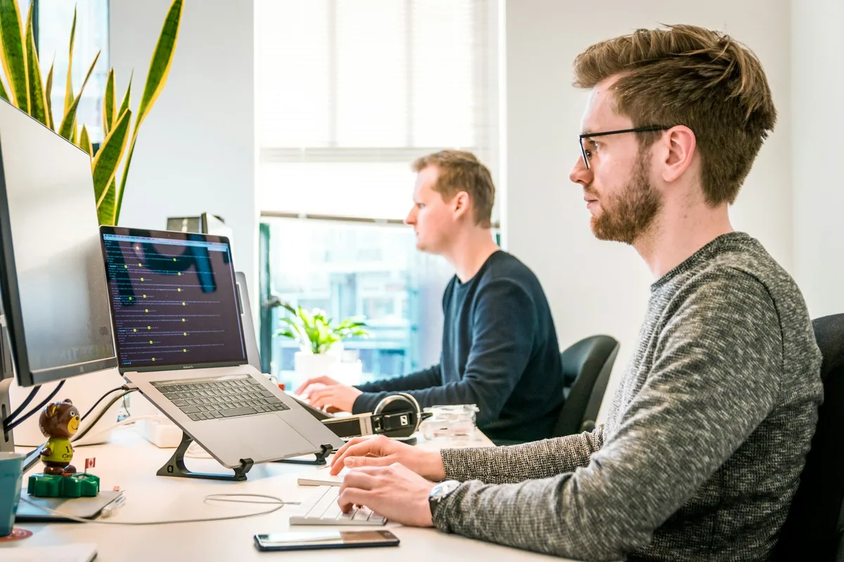 Two software professionals working at computers in a bright office, with code on a laptop screen.