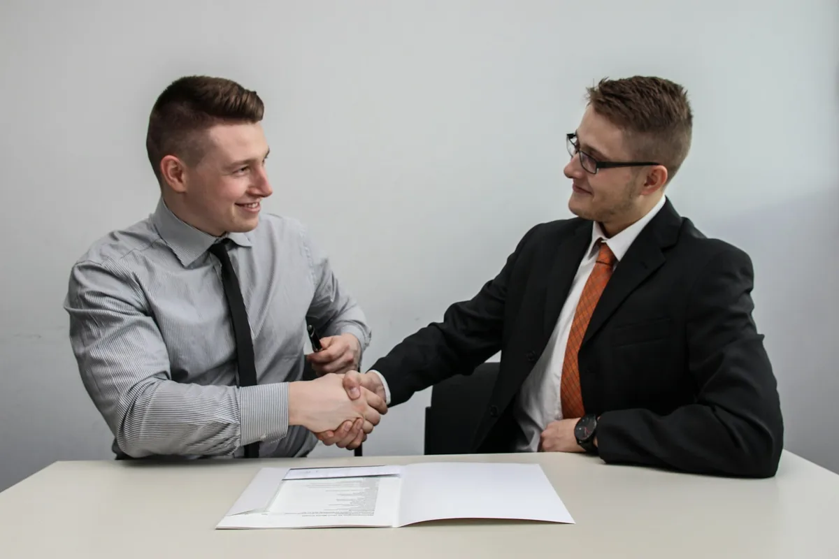 Two professionals smile and shake hands across a desk during a business meeting